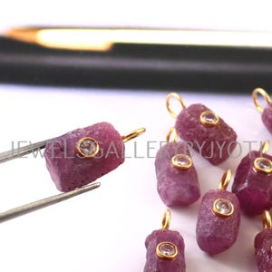 May include: Close-up of several rough-cut, deep pink ruby pendants with gold-colored loops and small, clear, round accents. One pendant is held by tweezers. The pendants are on a white surface.