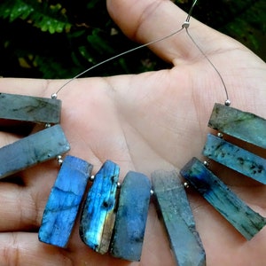 May include: A strand of labradorite gemstone beads, each bead is a rectangular shape with a blue, green, and gold iridescent sheen. The beads are strung on a silver wire.