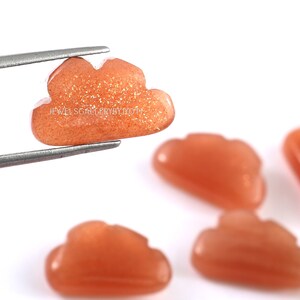 May include: Five orange sunstone cabochons carved in the shape of clouds. The cabochons are approximately 2.5 cm in size and have a sparkly, glittery finish.