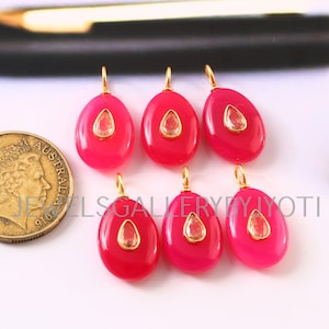 Peut inclure: Six pendentifs ovales rose vif avec des bélières dorées et une pierre transparente en forme de goutte au centre. Une pièce d'or est dans le coin inférieur gauche. Les pendentifs sont sur une surface blanche.
