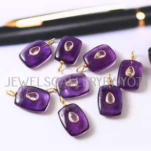May include: Close-up of several rectangular, purple amethyst pendants with gold-colored loops. Each pendant features a smaller, oval-shaped, clear stone in the center. The text "JEWELS GALLERY BY JYOTI" is partially visible.
