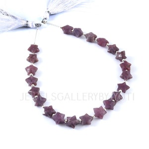 Puede incluir: Una hebra de cuentas de piedra preciosa en forma de estrella de un tono púrpura intenso. Las cuentas están enhebradas en un alambre de plata con separadores de plata entre cada cuenta. La hebra está terminada con dos borlas blancas.