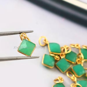 May include: Close-up of a collection of gold pendants, each featuring a square, light green gemstone. One pendant is held by tweezers. The pendants have a small loop at the top for hanging. The text "JEWELSGEMS BYJYOTI" is visible.