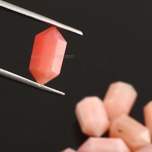 May include: A close-up of a pink, faceted gemstone held by tweezers. The gemstone is a hexagonal shape with a smooth, polished surface.  The gemstone is surrounded by other similar gemstones.
