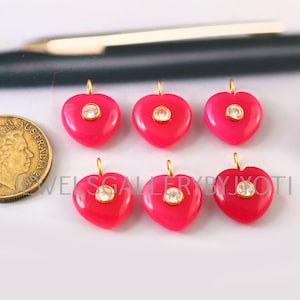 May include: Six bright pink heart-shaped pendants, each with a small, clear gemstone and a gold-coloured loop. A coin is visible on the left. The text "JEWELSGALLERYBYJYOTI" is partially visible.