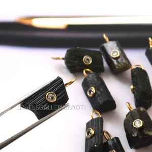 Może przedstawiać: Zbliżenie kilku ciemnozielonych i czarnych wisiorków z turmalinu ze złotymi krawędziami. Każdy wisiorek posiada mały, okrągły, przezroczysty kamień osadzony w złotej oprawie. Jeden wisiorek jest trzymany przez srebrną pęsetę. Widoczny jest tekst "JEWELSGALLERYJYOTI".