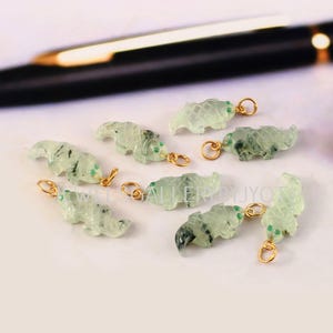 Puede incluir: Un conjunto de pequeños dijes de animales tallados en color verde pálido translúcido, cada uno con una anilla de metal dorado en la parte superior. Los dijes presentan una superficie texturizada y escamada con inclusiones oscuras.