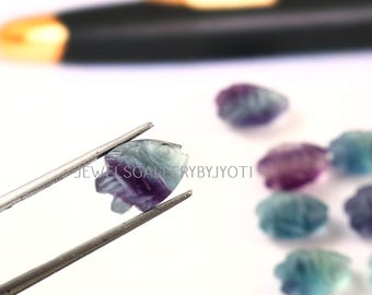 Puede incluir: Un pequeño pez de fluorita tallado, sostenido por unas pinzas, que muestra tonos de púrpura, azul y verde. El pez tiene detalles tallados. Otros peces similares están en el fondo. El texto "JEWELSGALLERYBYJYOTI" es visible.