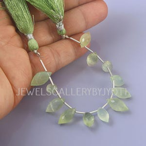 May include: A strand of pale green gemstone beads in a leaf shape, strung on a silver wire. The beads are held by a hand, with two green tassel accents at the top. The background is a light purple color. The text "JEWELSGALLERYBYJYOTH" is visible.