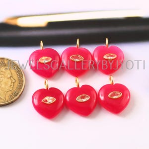 May include: Six red heart-shaped pendants, each with a gold-coloured loop and a clear, oval-shaped stone. The pendants are displayed on a white surface. A gold coin and a gold pen are visible in the background. The pendants are likely for jewellery making.