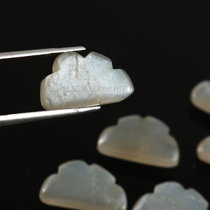 Pode incluir: Cinco cabochões de pedra da lua branca esculpidos em forma de nuvem. Os cabochões são lisos e polidos, com um leve brilho iridescente.