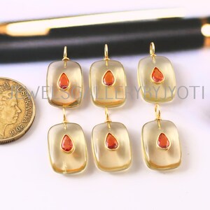 May include: Six gold-toned pendant earrings with rectangular, translucent centers and teardrop-shaped, orange-red gemstones. The earrings are arranged in two rows of three, with a coin visible on the left.