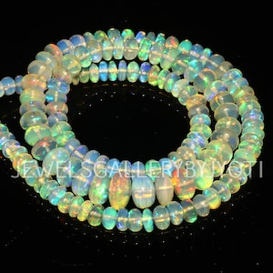May include: Strands of opalescent opal beads in various sizes. The beads display a range of colors, including white, yellow, green, and blue, with iridescent flashes. The text "JEWELSGALLERYBYJYOTI" is visible.