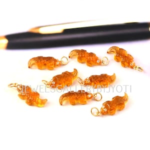 Peut inclure: Un ensemble de huit petites breloques en pierre gemme orange translucide sculptées en forme de crocodiles. Chaque breloque présente une surface facettée et un petit anneau de jonction en métal doré fixé au sommet pour servir de pendentif.