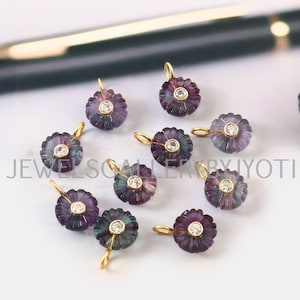 May include: A collection of flower-shaped pendants. Each pendant features a carved purple and green gemstone with a central clear stone and a gold-colored bail. The pendants are arranged on a white surface.