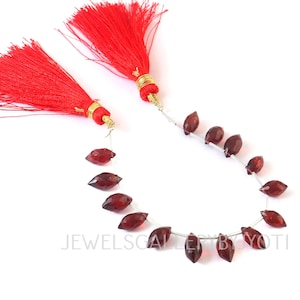 May include: Strands of dark red, faceted gemstone beads with a leaf shape, strung on silver wire. Two bright red tassels with gold accents are at the top. The beads are displayed against a white background.