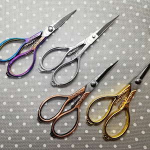 Pode incluir: Quatro pares de tesouras decorativas com alças ornamentadas. As tesouras são de diferentes cores: roxo, prata, cobre e dourado.