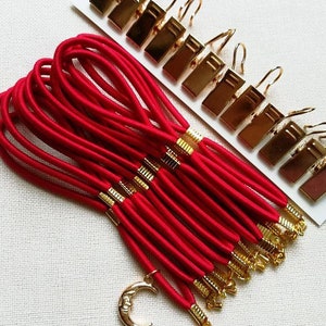Puede incluir: Un conjunto de cordón elástico rojo con puntas de metal dorado y un dije de media luna. El cordón está agrupado y hay 12 clips de metal dorado con ganchos en el lateral.