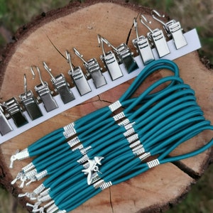 May include: A set of teal elastic cord with silver metal clips and a silver airplane charm. The cord is wrapped around a wooden log.