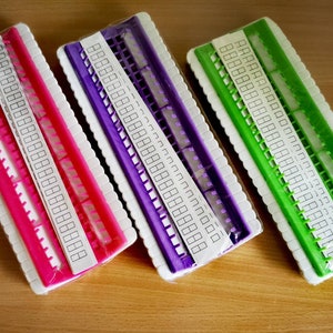 Puede incluir: Tres tableros de telar de plástico en rosa, morado y verde. Cada tablero tiene una etiqueta blanca con filas de números.