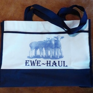 May include: A rectangular tote bag featuring a cream-colored main body with a dark blue bottom panel and matching dark blue handles. The front displays a blue graphic of three lambs and the text EWE~HAUL printed in a bold, dark blue font.