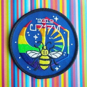 Può includere: Una toppa ricamata blu con una mezzaluna arcobaleno, un'ape con strisce gialle e nere e il testo "2019 UT17V".