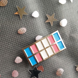 May include: A silver pin with a grid of nine squares. The squares are alternating colours of blue, pink, and white.