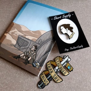 Può includere: Illustrazione di un personaggio di un film di fantascienza, seduto in un paesaggio desertico. Una spilla del profilo del personaggio è su una carta nera. È presente anche una spilla con la spada laser con la scritta "STRONGER THAN SHE KNOWS".