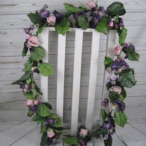 May include: A garland of artificial flowers with pink roses and purple flowers, with green leaves, draped over a white wooden fence.