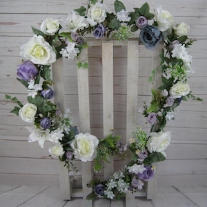 May include: A white wooden heart-shaped frame decorated with a garland of white, purple, and blue artificial flowers. The garland is made of roses, lilies, and other small flowers.