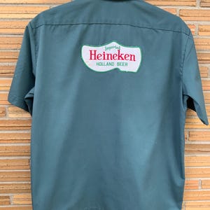 K&ouml;nnte beinhalten: Ein dunkelgr&uuml;nes Kurzarmhemd mit einem gro&szlig;en wei&szlig;en Aufn&auml;her auf der R&uuml;ckseite. Der Aufn&auml;her ist in Form eines Holzschuhs gestaltet und zeigt den roten Schriftzug Heineken sowie die gr&uuml;nen W&ouml;rter Imported und HOLLAND BEER.
