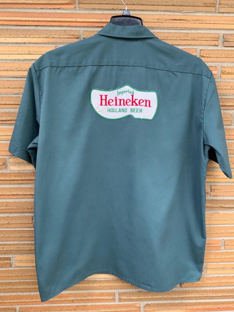 K&ouml;nnte beinhalten: Ein dunkelgr&uuml;nes Kurzarmhemd mit einem gro&szlig;en wei&szlig;en Aufn&auml;her auf der R&uuml;ckseite. Der Aufn&auml;her ist in Form eines Holzschuhs gestaltet und zeigt den roten Schriftzug Heineken sowie die gr&uuml;nen W&ouml;rter Imported und HOLLAND BEER.