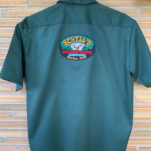 May include: A dark green short-sleeved button-down shirt featuring an oval patch on the back. The patch displays a deer head illustration, the text "SCHELL'S", "IT'S A GRAND OLD BEER", "SINCE 1860", and "NEW ULM, MINN" in yellow, red, and blue.