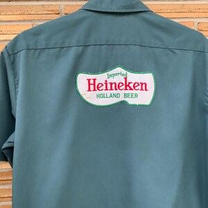 K&ouml;nnte beinhalten: Ein kurz&auml;rmeliges, blaugr&uuml;nes Hemd mit einem gro&szlig;en, wei&szlig;en Aufn&auml;her auf der R&uuml;ckseite, der wie ein Holzschuh geformt ist. Der Aufn&auml;her zeigt den roten Schriftzug Heineken sowie die gr&uuml;nen W&ouml;rter Imported und HOLLAND BEER &uuml;ber und unter dem Namen.