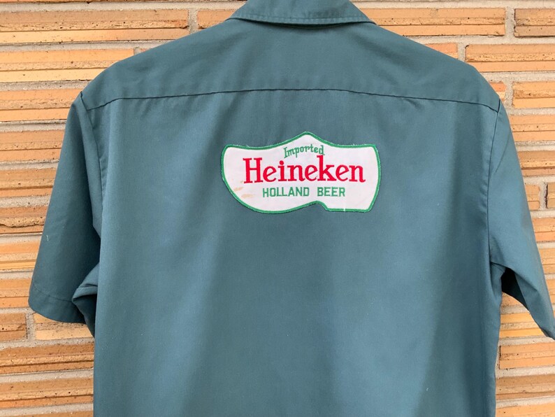 K&ouml;nnte beinhalten: Ein kurz&auml;rmeliges, blaugr&uuml;nes Hemd mit einem gro&szlig;en, wei&szlig;en Aufn&auml;her auf der R&uuml;ckseite, der wie ein Holzschuh geformt ist. Der Aufn&auml;her zeigt den roten Schriftzug Heineken sowie die gr&uuml;nen W&ouml;rter Imported und HOLLAND BEER &uuml;ber und unter dem Namen.