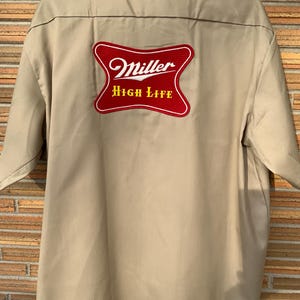 May include: Tan work shirt with a red and white Miller High Life patch on the back. The shirt has a button-down collar and short sleeves. The patch features the Miller logo and the words "High Life".