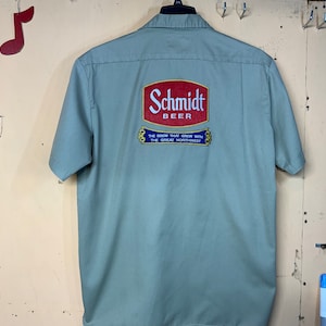 Könnte beinhalten: Ein hellgrünes Arbeitshemd mit einem roten und weißen gestickten Logo, auf dem "Schmidt Beer" und "The Brew That Grew With The Great Northwest" steht.