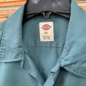 K&ouml;nnte beinhalten: Ein blaugr&uuml;nes Dickies-Arbeitshemd mit spitzem Kragen und Knopfleiste. Das innere Nackenetikett zeigt das Dickies-Logo, die Gr&ouml;&szlig;e XL und die Materialzusammensetzung von 65% Polyester und 35% Baumwolle, hergestellt in Honduras.