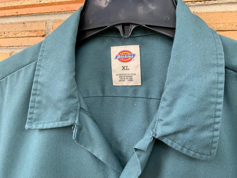 K&ouml;nnte beinhalten: Ein blaugr&uuml;nes Dickies-Arbeitshemd mit spitzem Kragen und Knopfleiste. Das innere Nackenetikett zeigt das Dickies-Logo, die Gr&ouml;&szlig;e XL und die Materialzusammensetzung von 65% Polyester und 35% Baumwolle, hergestellt in Honduras.