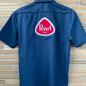 May include: Navy blue short-sleeved work shirt with a button-down collar. The back features a white and red logo that reads "Pearl Beer". The shirt is hanging on a black hanger.