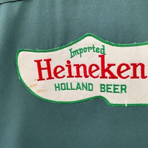 K&ouml;nnte beinhalten: Ein dunkelgr&uuml;nes Kleidungsst&uuml;ck aus Stoff mit einem wei&szlig;en, holzschuhf&ouml;rmigen Aufn&auml;her mit gr&uuml;nem Rand. Der Aufn&auml;her zeigt den Schriftzug "Imported" in Gr&uuml;n, "Heineken" in gro&szlig;en roten Buchstaben und "HOLLAND BEER" in gr&uuml;nem Text unten.