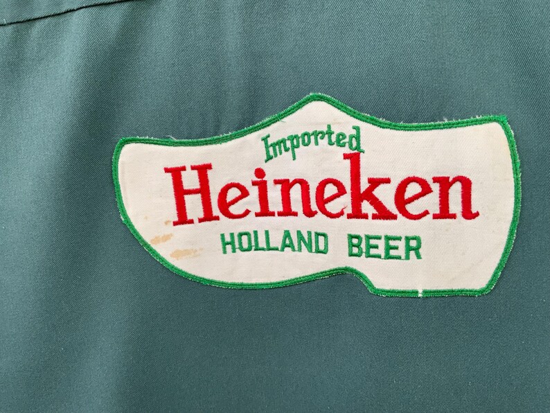 K&ouml;nnte beinhalten: Ein dunkelgr&uuml;nes Kleidungsst&uuml;ck aus Stoff mit einem wei&szlig;en, holzschuhf&ouml;rmigen Aufn&auml;her mit gr&uuml;nem Rand. Der Aufn&auml;her zeigt den Schriftzug "Imported" in Gr&uuml;n, "Heineken" in gro&szlig;en roten Buchstaben und "HOLLAND BEER" in gr&uuml;nem Text unten.