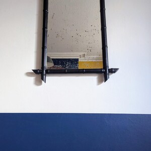 May include: A black framed mirror with a bamboo design hangs on a white wall with a blue accent wall below. The mirror has a chain hanging from the top.