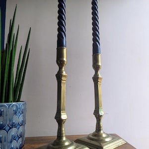 Op de afbeelding: Een paar gouden metalen kandelaars met vierkante bases en gedraaide zwarte kaarsen.