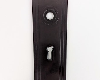 Könnte beinhalten: Schwarze Metall-Türplatte mit Art-Deco-Design. Sie hat ein Schlüsselloch und ein rundes Loch für einen Türknauf.