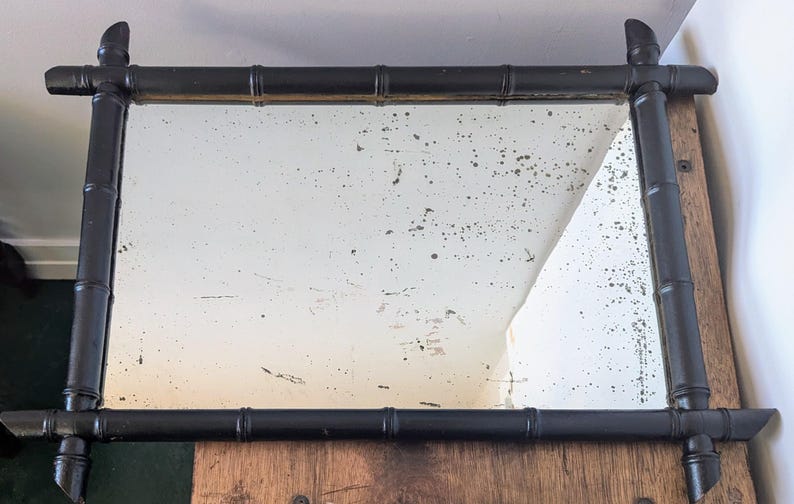 May include: A black framed mirror with a distressed, aged look. The frame is made of faux bamboo and has a rectangular shape.