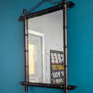 May include: A black bamboo framed mirror with a chain hanging from the top. The mirror is reflecting a white wall and a sign that says "FREE YOU DOOLY TOY".