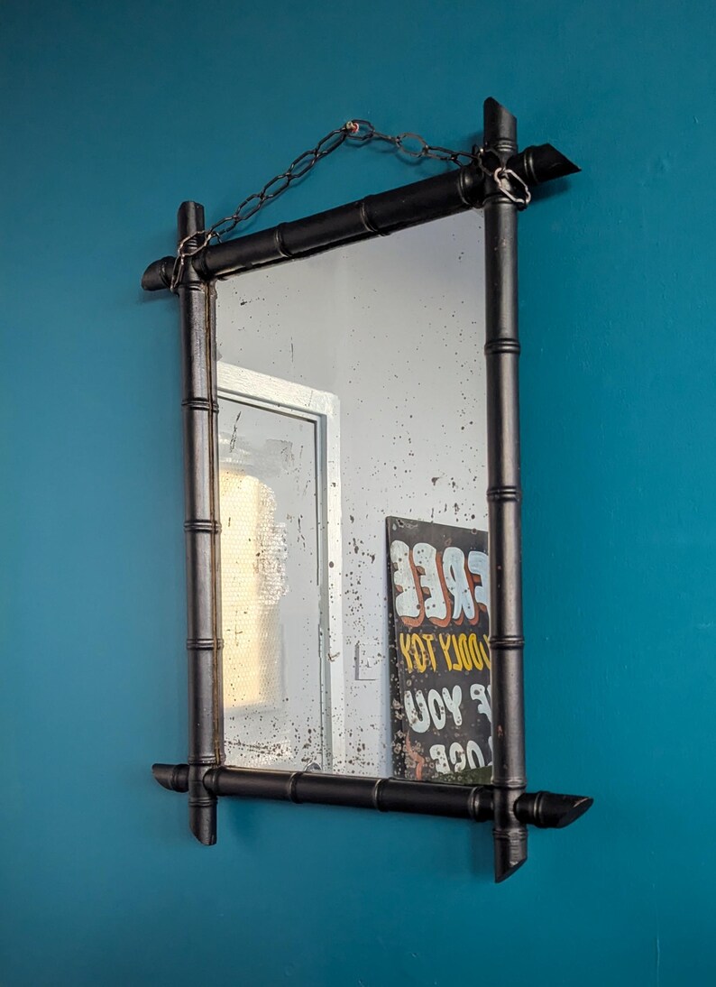 May include: A black bamboo framed mirror with a chain hanging from the top. The mirror is reflecting a white wall and a sign that says "FREE YOU DOOLY TOY".