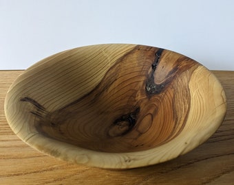 May include: A wooden bowl with a natural wood finish. The bowl has a light brown color with darker brown streaks throughout. The bowl is round and has a slight curve to the sides.