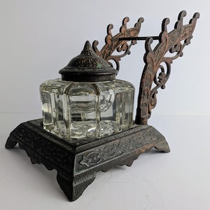 May include: Antique inkwell with a clear glass ink pot and a decorative metal base and pen rest. The ink pot has a faceted design and a matching lid. The metal base and pen rest have an ornate, aged appearance.